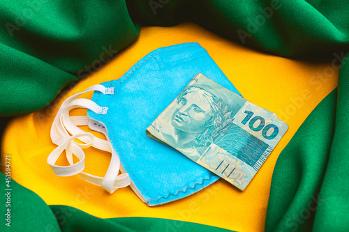 Real - BRL. A pandemia do novo coronavírus e economia brasileira. Uma cédula de 100 reais sobre uma máscara n95 de cor azul. Na composição da foto uma representação da bandeira do Brasil. photo