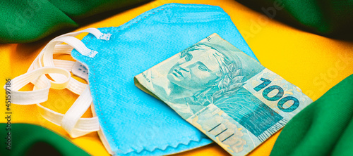  Real - BRL. A pandemia do novo coronavírus e economia brasileira. Uma cédula de 100 reais sobre uma máscara n95 de cor azul. Na composição da foto uma representação da bandeira do Brasil. photo