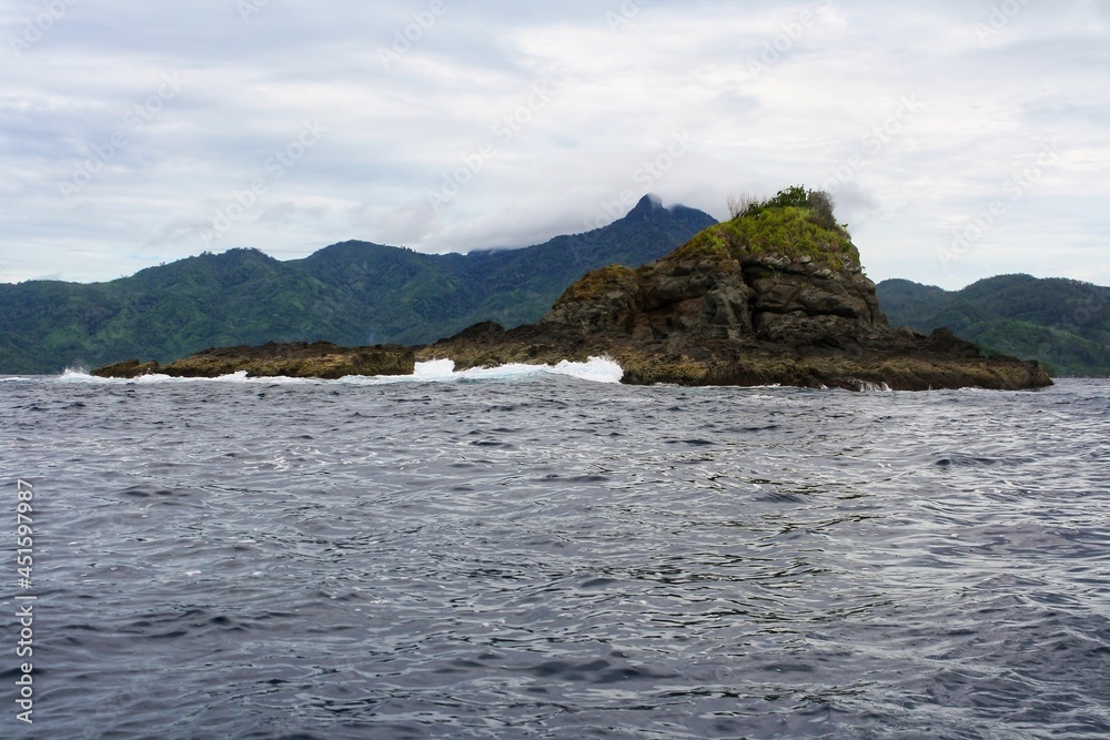 Scenic view of small tropical rock island in sea with dramatic clouds in blue sky and mountain in background. No people. Scenic view of small tropical rock island in sea with dramatic clouds in blue sky and mountain in background. No people.