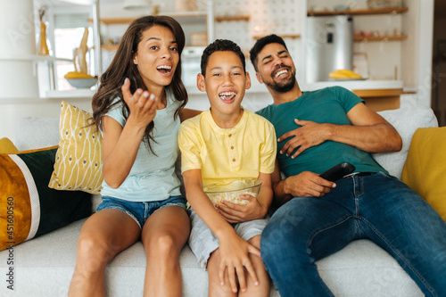 A happy mixed-race family is sitting on the couch at their home and watching comedy on television. The family is laughing and spending quality time together.