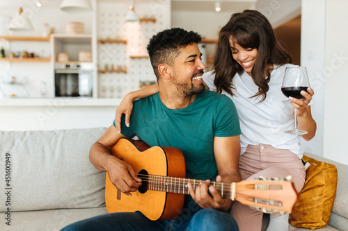 The African American couple is spending romantic moments together. The man is playing the guitar with his beloved girlfriend. A woman is sitting next to him on the couch and drinking red wine.
