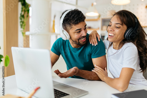 African couple is sitting at the dining table with headphones on and having a video call with their family or friends. They are so happy together, and they aren't hiding it. © Astarot