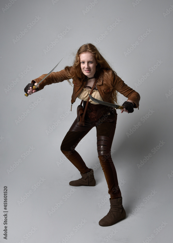 Full length  portrait of beautiful young woman with long red hair, wearing steampunk inspired costume knife weapons isolated on studio background.