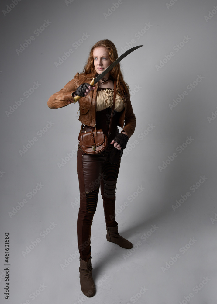 Full length portrait of beautiful young woman with long red hair, wearing steampunk inspired costume knife weapons isolated on studio background. Full length portrait of beautiful young woman with long red hair, wearing steampunk inspired costume knife weapons isolated on studio background.