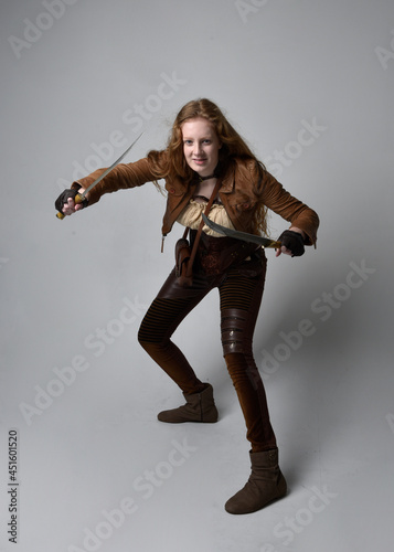 Full length portrait of beautiful young woman with long red hair, wearing steampunk inspired costume knife weapons isolated on studio background.