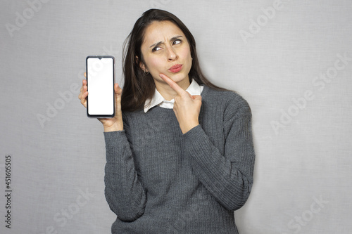 Mujer dudosa sosteniendo celular. Fondo blanco.