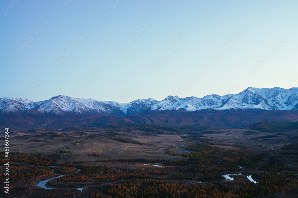 Awesome landscape with mountain river serpentine in valley among hills and forest in autumn colors with view to great snowy mountain range in sunset. High snow-covered mountains and autumn valley.