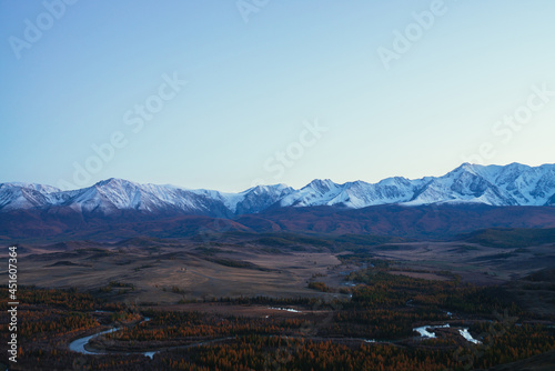 Awesome landscape with mountain river serpentine in valley among hills and forest in autumn colors with view to great snowy mountain range in sunset. High snow-covered mountains and autumn valley.