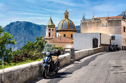 Romantic typical Italian street scene in the mountains of Positano on the Amalfi Coast in Italy with a motor scooter in front of Positano Cathedral, mountains and blue sky