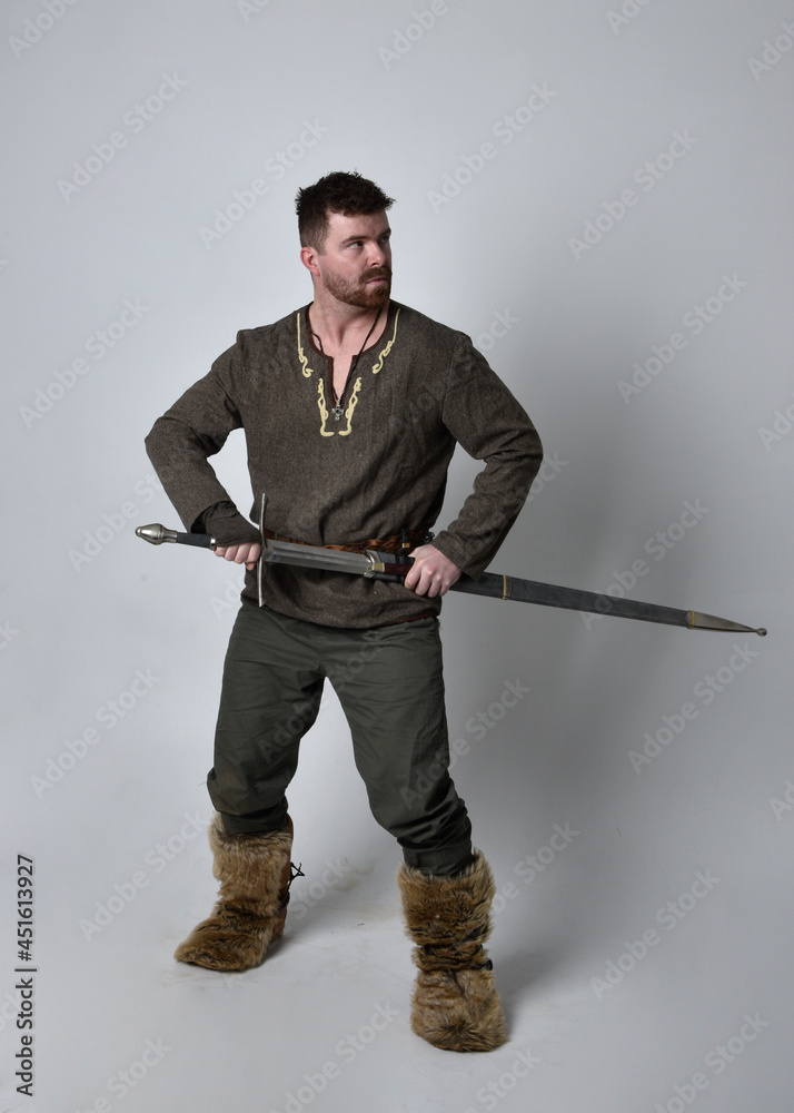 Full length portrait of young handsome man wearing medieval Celtic adventurer costume. Standing pose holding a sword, isolated on studio background. Full length portrait of young handsome man wearing medieval Celtic adventurer costume. Standing pose holding a sword, isolated on studio background.