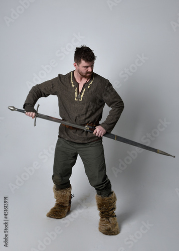 Full length portrait of young handsome man wearing medieval Celtic adventurer costume. Standing pose holding a sword, isolated on studio background.