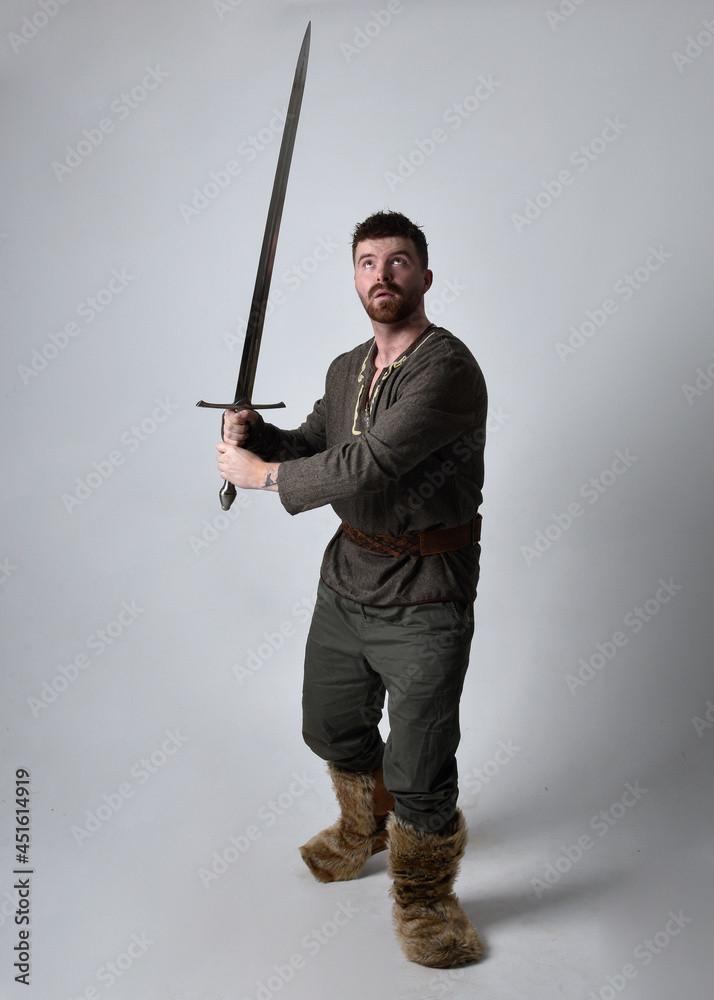 Full length portrait of young handsome man wearing medieval Celtic adventurer costume. Standing pose holding a sword, isolated on studio background. Full length portrait of young handsome man wearing medieval Celtic adventurer costume. Standing pose holding a sword, isolated on studio background.