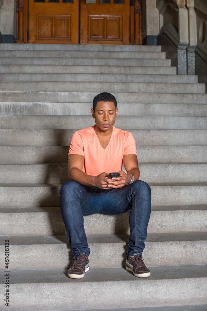 Dressing in a light orange short sleeve V neck shirt, jeans, a young handsome black student is sitting on stairs outside an office building, checking messages on his mobile phone.