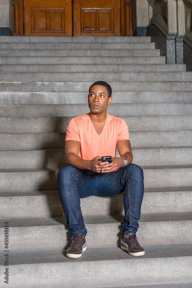 Dressing in a light orange short sleeve V neck shirt, jeans, a young handsome black student is sitting on stairs outside an office building, checking messages on his mobile phone
