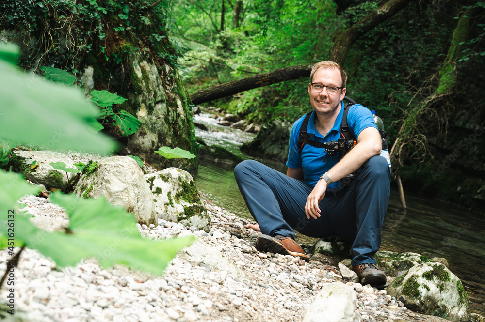 Smiling male photographer with backpack and camera sitting by a stream in the jungle