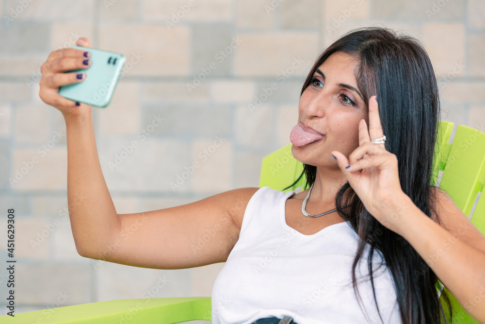 Young woman making silly face and showing tongue to smartphone while making selfie. Happy person having fun and making v sign with hand. Girl taking funny photo. Young woman making silly face and showing tongue to smartphone while making selfie. Happy person having fun and making v sign with hand. Girl taking funny photo.