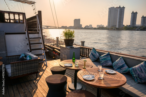 Restaurant on the deck with dining table, sofa, dishware, cutlery on ship in riverside at the evening