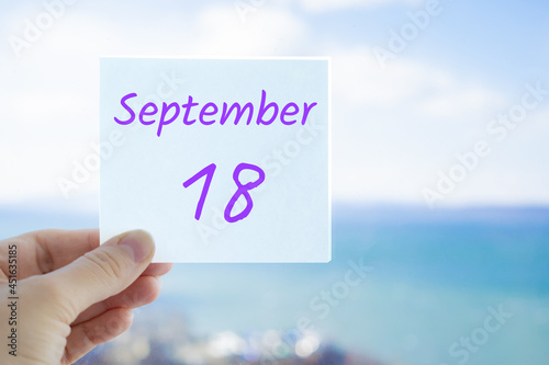 18th September. Hand holding sticker with text September 18 on the blurred background of the sea and sky. Copy space for text. Month in calendar concept