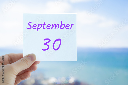 30th September. Hand holding sticker with text September 30 on the blurred background of the sea and sky. Copy space for text. Month in calendar concept