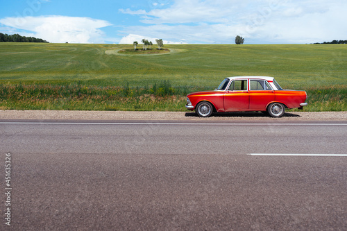 Red retro car is standing on the side of the road near an endless green field. Car trip among breathtaking beauties of nature