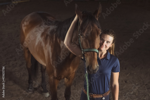 Horsewoman looking at her seal brown horse with love in the stable. Girl holding the stallion's rope with affection. Bonding between human beings and horses concept. Love for horses.
