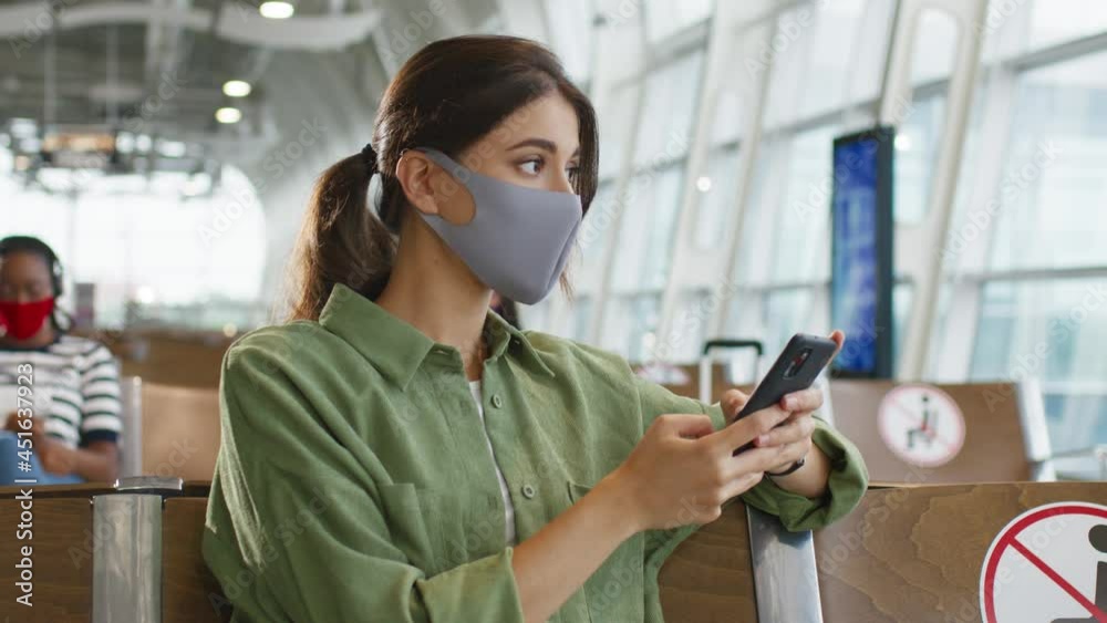 Charming young caucasian brown-haired girl wearing protective mask tapping and texting a message on the smartphone and sitting in the airport hall Charming young caucasian brown-haired girl wearing protective mask tapping and texting a message on the smartphone and sitting in the airport hall