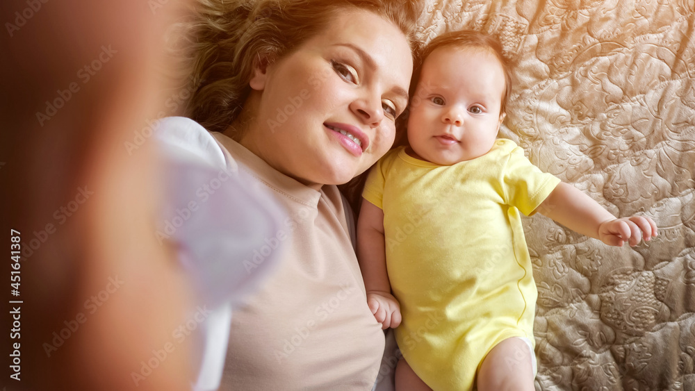 Jolly young mother with plump lips takes picture of smiling baby daughter in yellow with smartphone on bed in apartment room closeup Jolly young mother with plump lips takes picture of smiling baby daughter in yellow with smartphone on bed in apartment room closeup