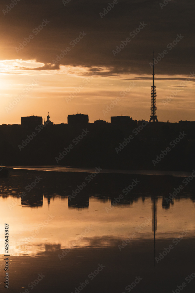 Golden epic contrast summer sunrise view in city vertical landscape with reflection in lake mirror water surface. Oleksiivske Reservoir and TV tower silhouette, Oleksiivka district in Kharkiv, Ukraine