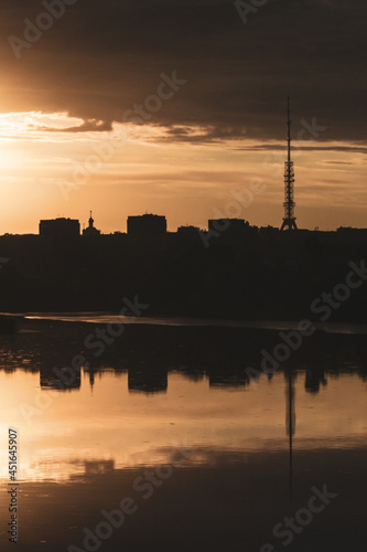 Golden epic contrast summer sunrise view in city vertical landscape with reflection in lake mirror water surface. Oleksiivske Reservoir and TV tower silhouette, Oleksiivka district in Kharkiv, Ukraine