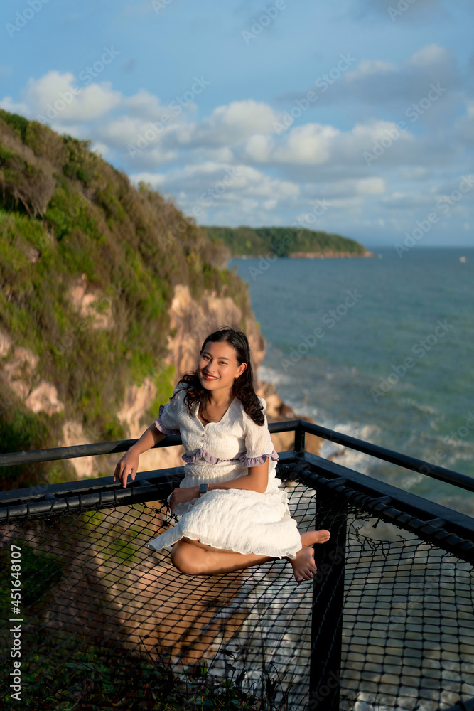 Asian woman tourist sit at net terrace on top hill with view above cliffs and sea in evening with sunlight, sky view, sunshine viewpoint in Phasuk nirun, Ko Proet, in Chanthaburi, Thailand