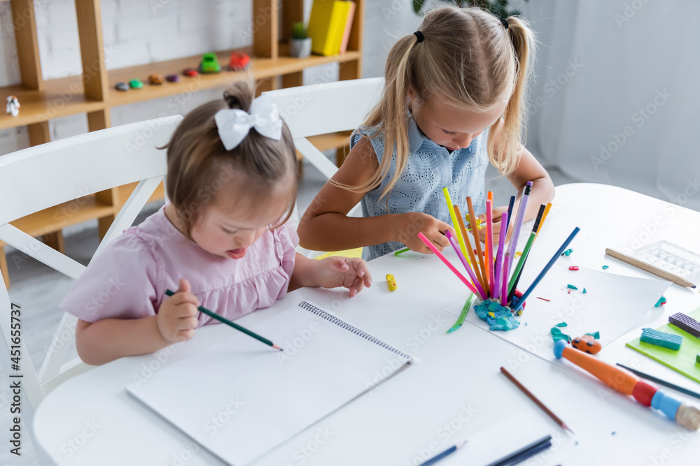 high angle view of girl with down syndrome drawing on paper near child in playroom high angle view of girl with down syndrome drawing on paper near child in playroom