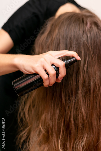 Young woman hand holds bottle and spraying her wavy blond long hair. Hair care for making it shiny, silken and smooth.