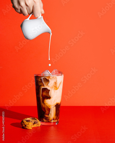 Iced coffee on a bright red background with a hand pouring cream into the coffee