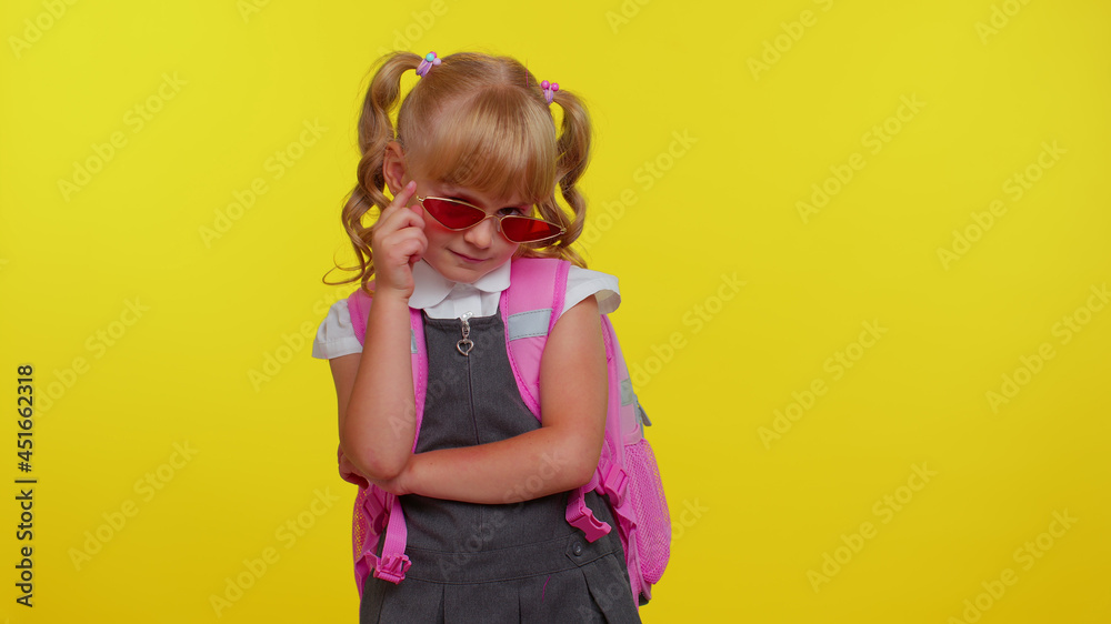 Portrait of joyful little smiling schoolgirl with backpack wearing sunglasses, charming smile, posing, dancing isolated over yellow studio background wall with copy space. Back to school concept