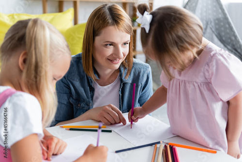 smiling kindergarten teacher looking at blurred toddler kid with down syndrome drawing with preschooler girl