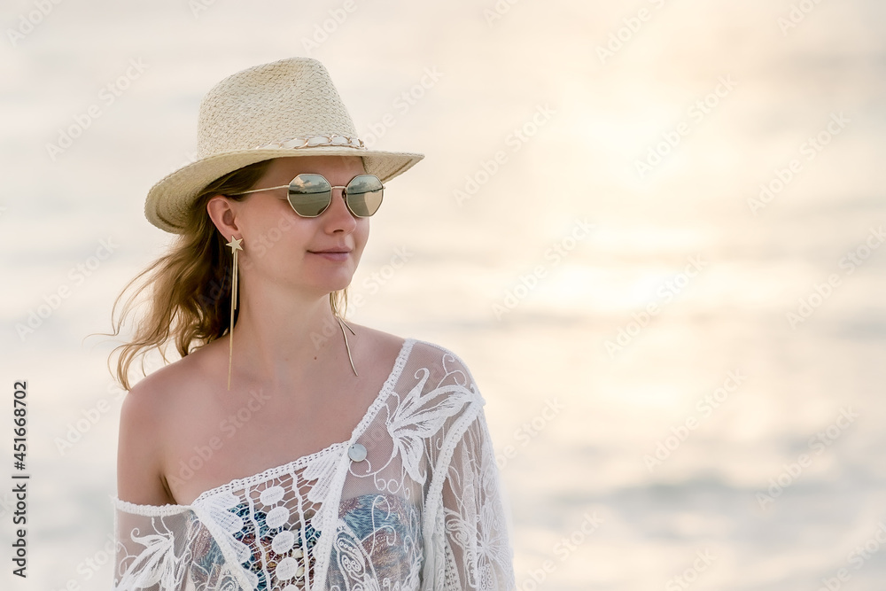 A portrait of an attractive 30 years old woman in the hat, sunglasses and long earrings posing on the beach