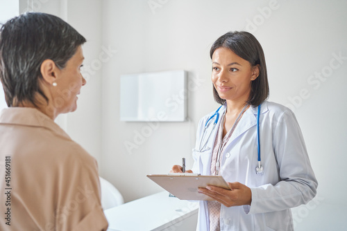 Doc at work. Young smiling african woman doctor in white coat with clipboard in hands feeling medical history, discussing symptoms with mature female patient visiting private clinic. Health checkup