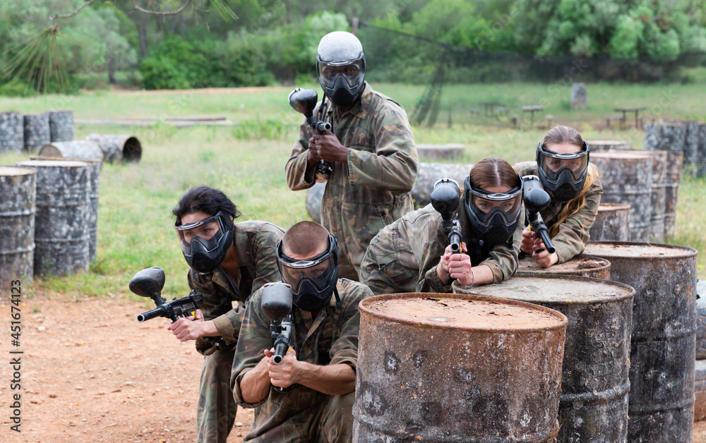 Fototapeta premium Five paintball players in camouflage and protective masks aims with guns outdoors
