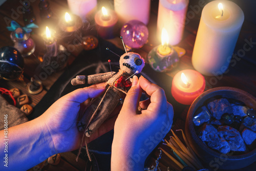 Sorceress or witch sticks needles into voodoo doll at ritual table with pentagram, burning candles and other occult objects, top view. Voodoo witchcraft, spirituality and occultism concept. photo