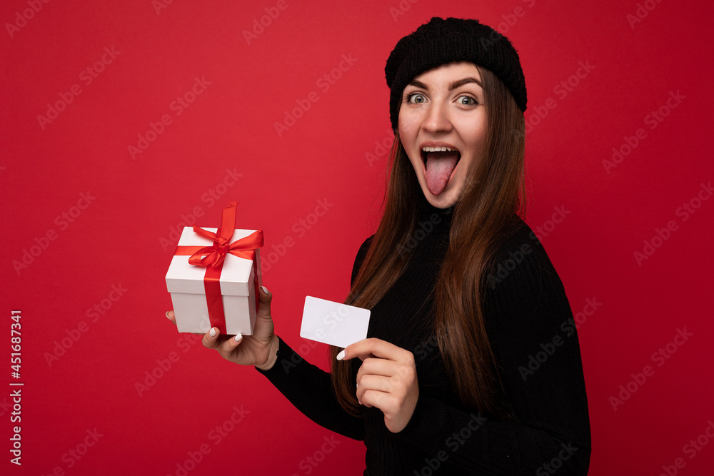 Beautiful happy funny young brunette woman wearing black sweater and hat isolated on red background holding credit card and gift box looking at camera and showing tongue