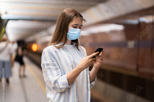 Young caucasian woman wear protective face mask, using mobile smart phone, chatting in social media, waiting for train on subway station platform during pandemic Covid-19. New normal concept. 