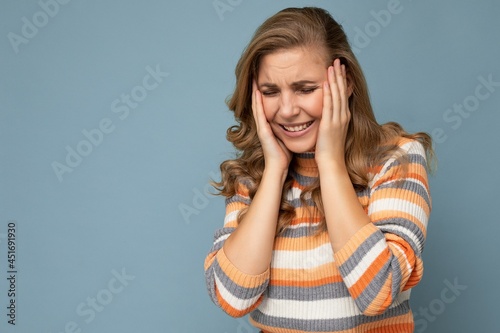 Portrait of young beautiful blonde curly woman with sincere emotions wearing stylish striped sweater isolated on blue background with empty space and having headache. Pain concept