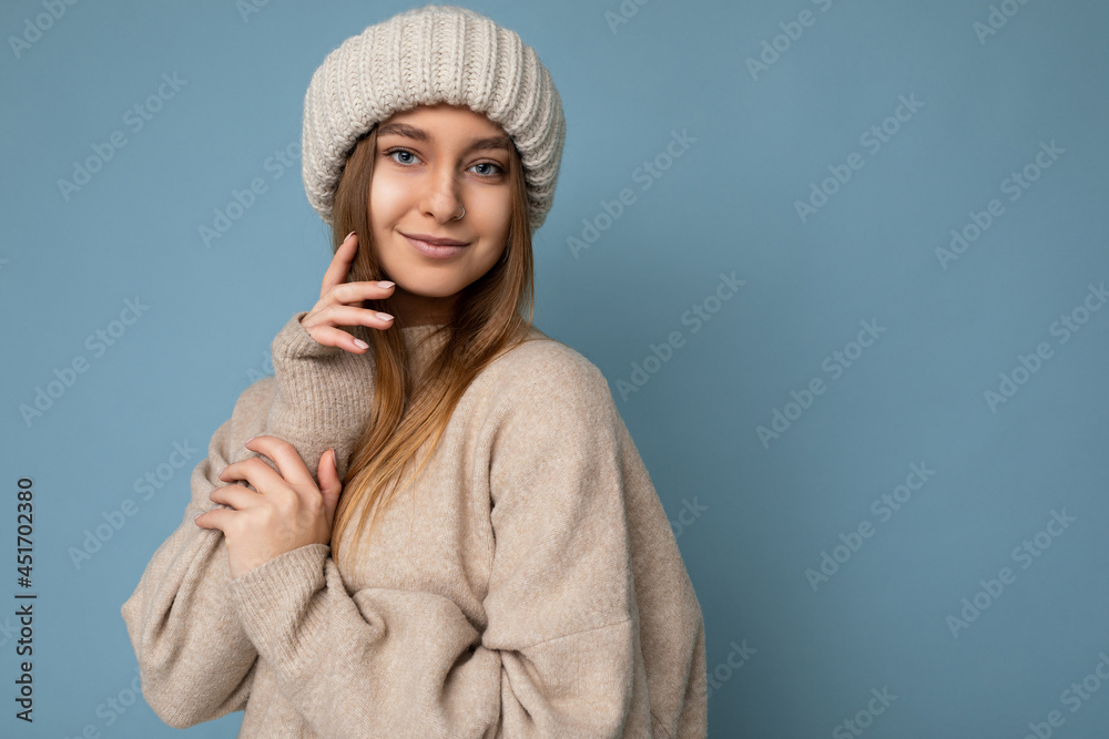 Shot of attractive happy cute sexy young dark blonde woman standing isolated on blue background wall wearing beige warm sweater and winter beige hat looking at camera Shot of attractive happy cute sexy young dark blonde woman standing isolated on blue background wall wearing beige warm sweater and winter beige hat looking at camera