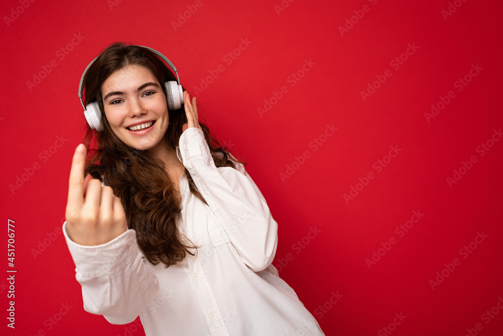 Attractive happy smiling young brunette female person wearing white shirt and optical glasses isolated over red background wearing white wireless bluetooth earphones listening to music and dancing Attractive happy smiling young brunette female person wearing white shirt and optical glasses isolated over red background wearing white wireless bluetooth earphones listening to music and dancing