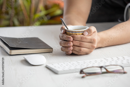 hands of a person holding a cup with hot coffee on a wooden desk, around a notebook, glasses, computer mouse, and keyboard. Office items photo