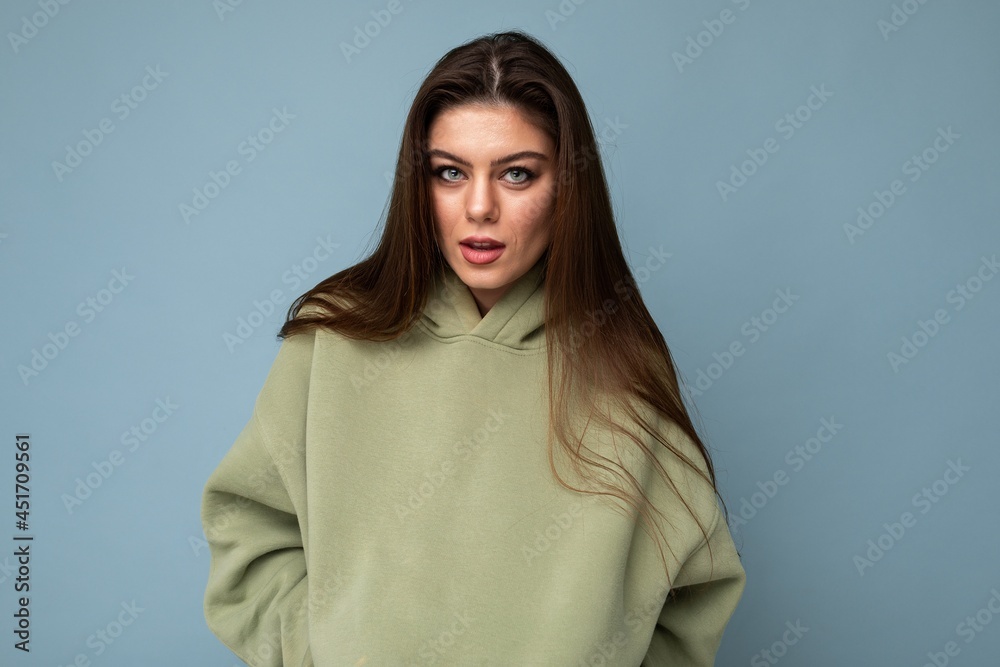 Shot of charming young brunet cool female person in a stylish khaki hoodie isolated on blue background. Free space Shot of charming young brunet cool female person in a stylish khaki hoodie isolated on blue background. Free space