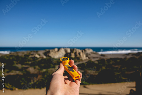 White hand holding a Yellow pipe with Marijuana  cannabis  in front of the ocean and the beach with rocks