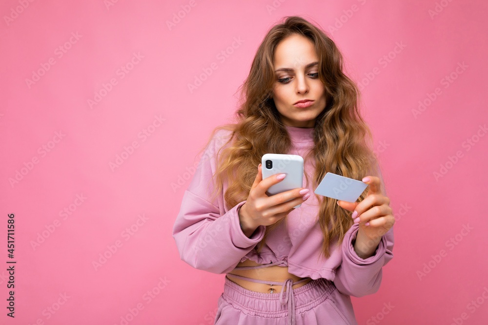 Beautiful young blonde curly woman wearing pink clothes isolated over pink background using mobile phone paying online shopping through credit card looking at smartphone display Beautiful young blonde curly woman wearing pink clothes isolated over pink background using mobile phone paying online shopping through credit card looking at smartphone display