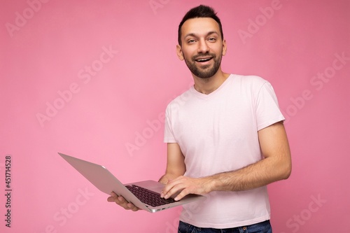 Handsome smiling brunet man holding laptop computer looking at camera in t-shirt on isolated pink background