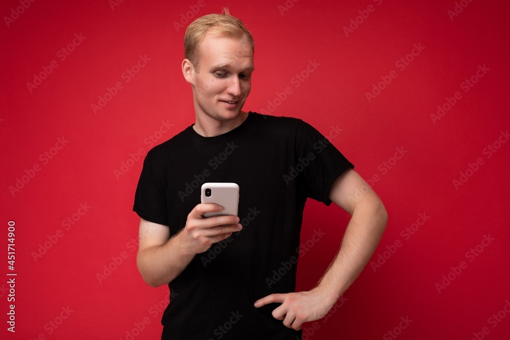 Photo of handsome smiling positive young blonde man isolated over red background wall wearing black t-shirt holding and using mobile phone writing sms looking at camera Photo of handsome smiling positive young blonde man isolated over red background wall wearing black t-shirt holding and using mobile phone writing sms looking at camera
